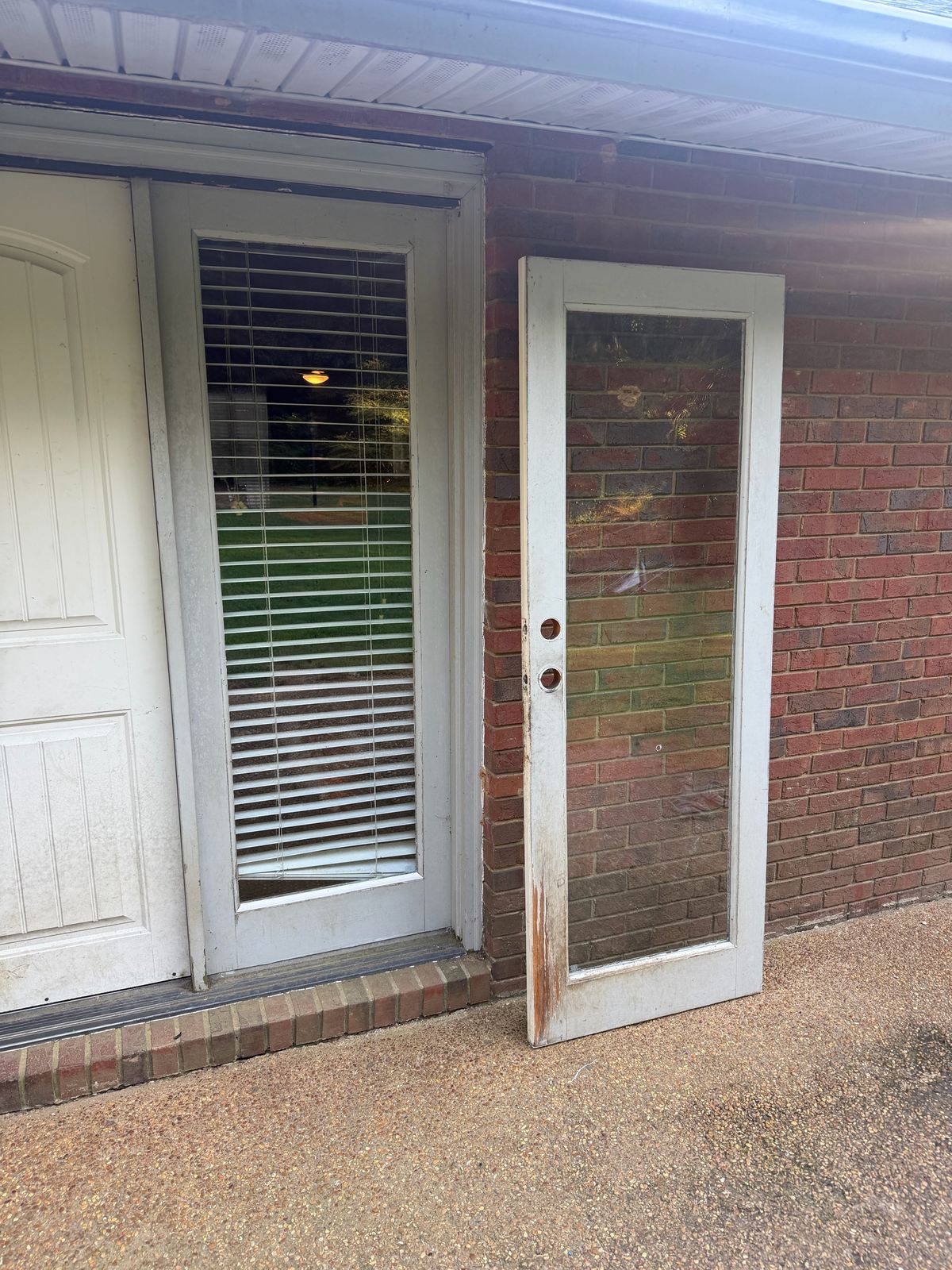 New full-view glass exterior door with blinds, next to the old glass door panel leaning on the brick wall